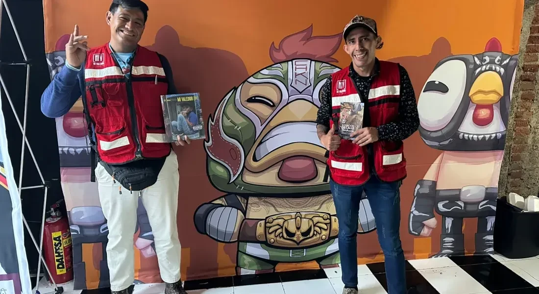 Dos personas con chalecos rojos posando frente a un mural de personajes en una feria
