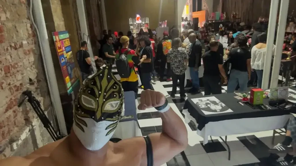Luchador enmascarado posando frente a una feria llena de visitantes y mesas de expositores